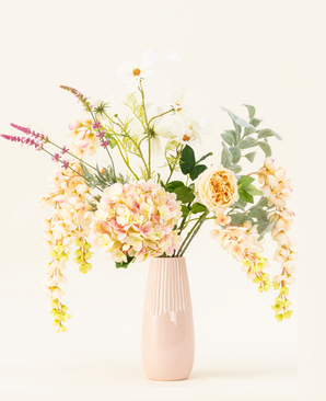 Een pastelroze vaas van keramiek toont het Zijden kunstbloemen boeket Blushing Beauty, met zachtgekleurde hortensia's, rozen en divers groen, prachtig gerangschikt tegen een bleke neutrale achtergrond.