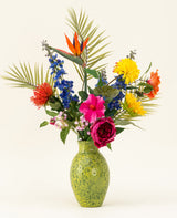 Een groene, gespikkelde vaas toont het Zijden kunstbloemen boeket Hibiscus Fiesta - met roze, rode en gele bloemen, blauwe riddersporen, oranje paradijsvogels en groene palmbladeren - tegen een effen lichte achtergrond.