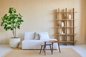 Salon minimaliste avec un canapé blanc, deux tables basses imbriquées en bois, des murs beiges, une étagère ouverte en bois avec des vases blancs et une Plante Artificielle Ficus 180 cm qui ajoute de la verdure. Un tapis beige recouvre le sol.