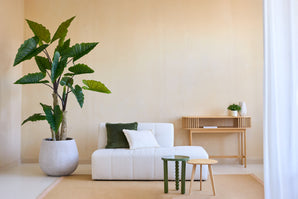 Un salon minimaliste avec la plante Artificielle Calocasia 210 cm, un canapé texturé blanc avec des coussins verts et blancs, deux petites tables d'appoint, un tapis beige et une console en bois contre un mur beige clair.