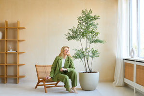 Une femme en costume vert sourit alors qu'elle est assise sur une chaise en bois à côté de la plante artificielle Ruscus de 180 cm dans une pièce lumineuse et moderne avec des murs beiges, de grandes fenêtres et une décoration minimaliste.