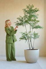Une femme en costume vert se tient à l'intérieur et sourit en touchant doucement les feuilles de la plante artificielle Ruscus de 180 cm. Un mur uni et de couleur claire sert de décor et crée une atmosphère calme et naturelle.