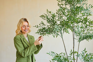 Une femme souriante vêtue d'un blazer vert se tient à l'intérieur et touche doucement les feuilles d'une plante artificielle de Ruscus de 150 cm avec de fines branches et un feuillage vert luxuriant, se détachant sur un fond uni de couleur claire.