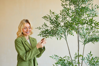Une femme souriante vêtue d'un blazer vert se tient à l'intérieur et touche doucement les feuilles d'une plante artificielle de Ruscus de 150 cm avec de fines branches et un feuillage vert luxuriant, se détachant sur un fond uni de couleur claire.