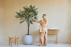 Une femme vêtue de vêtements amples beiges se tient à côté d’un olivier artificiel de 200 cm et touche doucement les feuilles. A proximité, un tabouret en bois et une console avec des livres et un diffuseur se trouvent contre le mur beige.