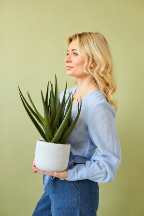 Une femme aux longs cheveux blonds vêtue d'un chemisier bleu clair et d'un pantalon bleu tient la plante artificielle Aloe Vera de 43 cm dans un pot sur un fond uni vert clair.