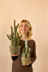 Une femme souriante vêtue d'une robe marron tient deux plantes en pot artificielles Cactus Cereus de 35 cm, une grande et une petite, sur un fond beige uni.