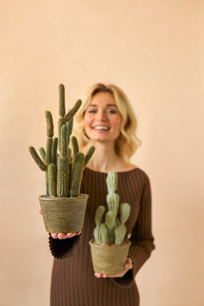 Une femme souriante vêtue d'une robe marron tient deux plantes en pot artificielles Cactus Cereus de 35 cm, une grande et une petite, sur un fond beige uni.