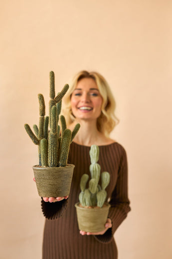 Une femme souriante vêtue d'une robe marron tient deux plantes en pot artificielles Cactus Cereus de 35 cm, une grande et une petite, sur un fond beige uni.