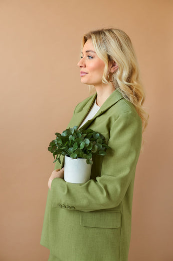 Une femme aux longs cheveux blonds vêtue d'un blazer vert clair tient la "Plante succulente artificielle 22 cm en pot décoratif" contre sa poitrine et se tient de côté devant un fond uni beige.