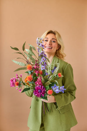 Une personne blonde souriante vêtue d'un costume vert tient le bouquet de fleurs artificielles en soie colorée Svenski sur un fond uni marron clair.