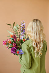 Une personne aux longs cheveux blonds ondulés et à la veste verte est vue de dos, tenant le bouquet de fleurs artificielles en soie Colorful Svenski sur un fond beige uni.