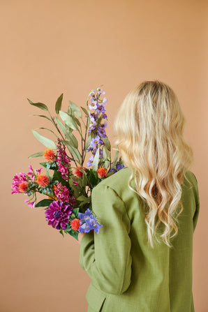 Une personne aux longs cheveux blonds ondulés et à la veste verte est vue de dos, tenant le bouquet de fleurs artificielles en soie Colorful Svenski sur un fond beige uni.