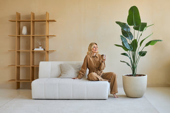 Une femme en tenue marron est assise sur un canapé blanc, souriant avec une tasse à la main. À côté d'elle se trouve la plante artificielle Strelitzia 180 cm real touch, qui apporte une verdure luxuriante dans le salon moderne aux tons beiges et au mobilier minimaliste.