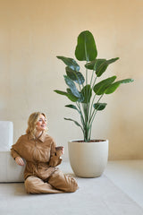 Une femme en combinaison marron est assise souriante sur le sol avec une tasse de café à côté d'elle, à côté de la plante artificielle Strelitzia 180 cm real touch dans une pièce minimaliste aux murs beiges.