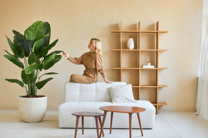 Une femme vêtue d'une combinaison marron est assise sur le dos d'un canapé blanc et touche la feuille d'une plante artificielle Strelitzia Real Touch Deluxe de 180 cm dans un salon minimaliste avec des tables et des étagères en bois.