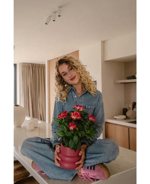 Une femme aux cheveux blonds bouclés, vêtue d'une combinaison en jean et de baskets roses, est assise les jambes croisées sur un comptoir de cuisine et sourit tout en tenant la plante artificielle Gardenia UV 39 cm de couleur fuchsia.