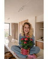 Une femme souriante aux cheveux blonds bouclés est assise les jambes croisées sur un comptoir lumineux et moderne, tenant la plante artificielle Gardenia UV fuchsia de 39 cm – une plante en pot rose avec des fleurs rouge vif. Elle porte un jean, une chemise bleue et des chaussures roses.