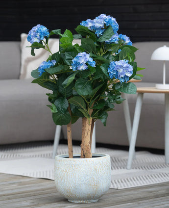 Une plante bleue d'hortensia artificielle UV de 85 cm avec des fleurs bleues et des feuilles vertes se dresse sur un parquet en bois devant un canapé gris et une petite table ronde avec une lampe blanche.