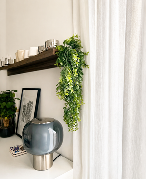 Un espace de vie moderne avec une lampe en verre gris, des œuvres d'art encadrées, une plante en pot et une étagère en bois avec des tasses et la plante artificielle suspendue « Pannenkoek » (61 cm), dont les vrilles pendent à côté de rideaux blancs.