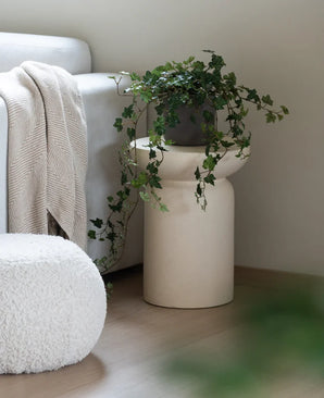 Un Lierre Artificiel Deluxe Vert 90 cm se dresse dans un vase beige sur un parquet en bois à côté d'un canapé gris clair avec un plaid de couleur crème et un pouf structuré blanc.