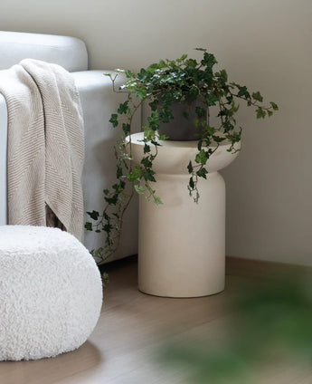Un Lierre Artificiel Deluxe Vert 90 cm se dresse dans un vase beige sur un parquet en bois à côté d'un canapé gris clair avec un plaid de couleur crème et un pouf structuré blanc.