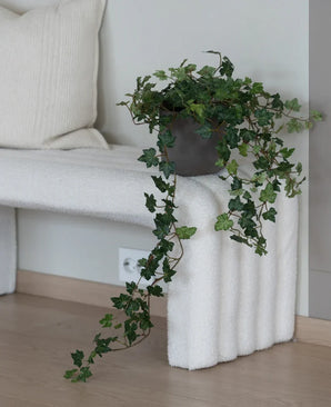 Le Lierre Artificiel Deluxe Vert 90 cm, avec ses vrilles suspendues luxuriantes, se dresse sur un banc structuré blanc à côté d'un coussin de couleur crème dans une pièce minimaliste au parquet en bois clair.