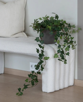 Le Lierre Artificiel Deluxe Vert 90 cm, avec ses vrilles suspendues luxuriantes, se dresse sur un banc structuré blanc à côté d'un coussin de couleur crème dans une pièce minimaliste au parquet en bois clair.
