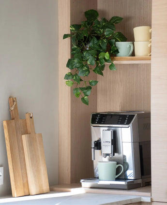 Sur le comptoir se trouve une cafetière en argent avec une tasse verte. Au-dessus, sur une étagère en bois, des tasses aux couleurs pastel sont empilées et un 'Art Hanging gold vine 60 cm UV' est suspendu. Juste à côté, des planches à découper en bois sont appuyées contre le mur.