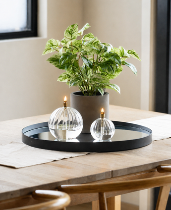 Une vigne dorée artificielle vert-blanc en pot UV 35 cm se tient avec deux lampes à huile en verre sur une plate-forme de miroir ronde sur une table en bois, éclairée par la lumière naturelle entrant à travers une fenêtre voisine dans cet environnement moderne et minimaliste.