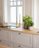 Une cuisine lumineuse avec des plans de travail en bois, des placards blancs, deux bougeoirs dorés, une poêle blanche sur la cuisinière et une grande fenêtre laissant entrer la lumière naturelle. L'espace est accentué par la Vigne Dorée Artificielle vert-blanc en pot UV 35 cm.