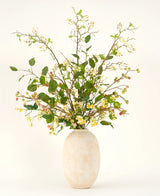 Un grand vase en céramique beige contient le bouquet de fleurs artificielles Silk Wild Apple Blossom XL, avec des branches feuillues vertes, de petites fleurs jaunes et des baies roses sur un fond clair.