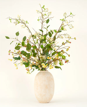 Un grand vase en céramique beige contient le bouquet de fleurs artificielles Silk Wild Apple Blossom XL, avec des branches feuillues vertes, de petites fleurs jaunes et des baies roses sur un fond clair.