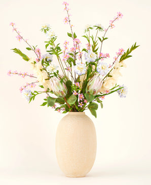 In een vaas met beige structuur staat het Zijden kunstbloemen boeket Lots of Love XL, een handgebonden boeket van roze, witte en blauwe zijden bloemen met groene bladeren en lange stelen tegen een lichte achtergrond.