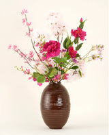 Le bouquet de fleurs artificielles en soie Pink Reverie se présente dans un vase à structure marron, avec des fleurs artificielles roses, magenta et blanches avec des feuilles vertes sur un fond uni clair.