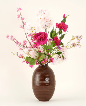 Le bouquet de fleurs artificielles en soie Pink Reverie se présente dans un vase à structure marron, avec des fleurs artificielles roses, magenta et blanches avec des feuilles vertes sur un fond uni clair.