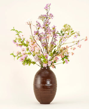 Un vase en céramique marron montre le bouquet de fleurs artificielles en soie Lilas, un bouquet de soie grand et élégant avec des fleurs roses, violettes et lilas avec des feuilles vertes et de fines branches sur un fond clair uni.
