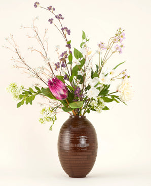 Un bouquet de fleurs artificielles en soie Serene Bloomfield, comprenant une grande protéa rose, des roses blanches, des fleurs violettes et blanches et un feuillage vert, se dresse dans un vase côtelé marron sur un fond clair uni.