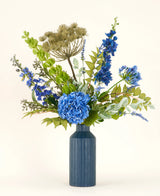 Le bouquet de fleurs artificielles en soie Mystic Blue Garden se compose d'hortensias bleus, de pied d'alouette, de feuilles vertes et de têtes de graines beiges séchées dans un vase en céramique bleue - idéal pour égayer votre espace sur un fond clair.