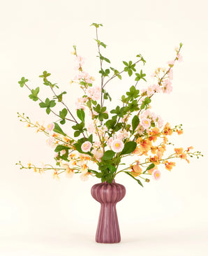 Un bouquet de fleurs artificielles en soie Blossom Harmony est disposé dans un vase mauve avec des fleurs roses et pêche et des feuilles vertes, sur un fond clair simple pour un affichage élégant.