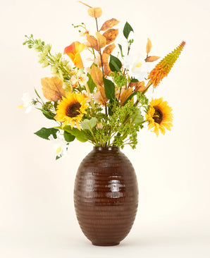 Un vase texturé marron montre le bouquet de fleurs artificielles en soie Amber Light, avec des tournesols, des fleurs blanches et oranges et un feuillage vert sur un fond uni.