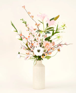 Un vase de couleur crème montre le bouquet de fleurs artificielles en soie Pink & Pearl, avec de délicates fleurs en soie roses, blanches et pêche avec des feuilles vertes sur un fond neutre clair.