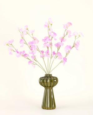 Un vase en verre vert montre le bouquet de fleurs artificielles en soie rose pois avec un feuillage vert tendre, avec plusieurs longues tiges et sur un fond uni blanc cassé.
