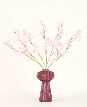 Le bouquet de fleurs artificielles en soie Pois rose clair, avec plusieurs tiges de pois rose clair dans un vase bordeaux côtelé, se dresse sur un fond pâle - idéal pour tout intérieur de fleurs artificielles.