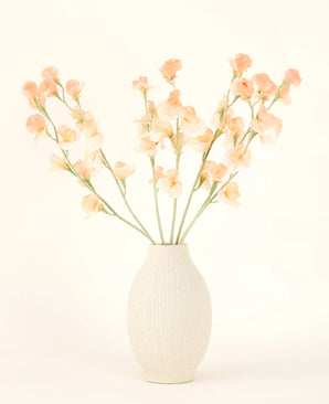 Le vase en céramique de couleur crème contient le bouquet de fleurs artificielles Silk Pea Peach, avec de délicates fleurs pêche et rose clair avec de longues tiges vertes sur un fond clair et neutre.