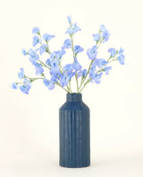 Un vase en céramique nervurée bleue présente le bouquet de fleurs artificielles en soie Pea Blue, avec ses délicates fleurs artificielles bleu clair sur un fond uni et clair.