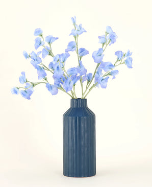 Un vase en céramique nervurée bleue présente le bouquet de fleurs artificielles en soie Pea Blue, avec ses délicates fleurs artificielles bleu clair sur un fond uni et clair.