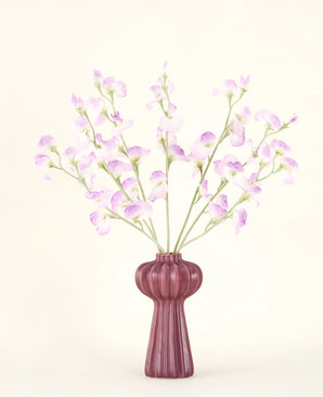 Le bouquet de fleurs artificielles en soie Pois lilas présente de longues tiges de fleurs délicates violet clair et de feuilles vertes dans un vase en céramique mauve aux stries verticales, sur un fond uni blanc cassé.