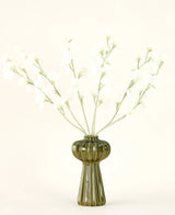Un vase en verre nervuré vert contient le bouquet de fleurs artificielles en soie Crème de pois avec de délicates fleurs en soie blanche et des tiges minces, sur un fond uni blanc cassé.