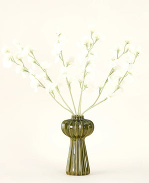 Un vase en verre nervuré vert contient le bouquet de fleurs artificielles en soie Crème de pois avec de délicates fleurs en soie blanche et des tiges minces, sur un fond uni blanc cassé.